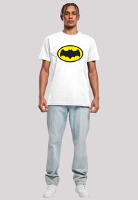 White cotton t-shirt featuring a large black and yellow Batman logo. Worn with light blue jeans and white sneakers, standing against a neutral background.