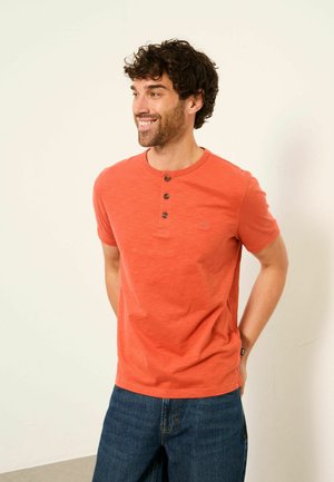 Lächelnder Mann mit lockigem Haar, der ein orangefarbenes Henley-Shirt mit kurzen Ärmeln und blaue Jeans trägt, steht mit den Händen hinter dem Rücken.