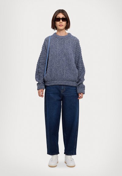 Suéter de punto azul grueso con hombros caídos, combinado con jeans anchos azules y zapatillas blancas, que presenta un diseño estructurado y un acabado texturizado.