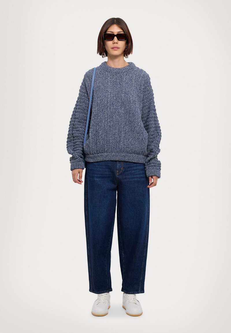 Chunky blauwe gebreide trui met verlaagde schouders, gecombineerd met losse blauwe jeans en witte sneakers, met een gestructureerd ontwerp en een textuurafwerking.