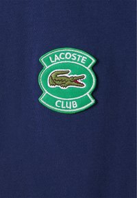 Navy blue cotton fabric with a green embroidered oval badge reading "LACOSTE" and "CLUB," featuring a crocodile design in green and red.