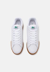 Bijele tenisice s glatkim sintetičkim gornjim dijelom, okruglim nosom, dizajnom s vezicama i smeškim gumenim potplatom. Lacoste logo prikazan na jeziku.