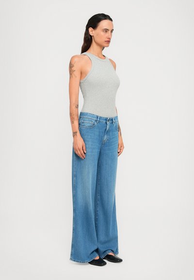 Woman with long dark hair wearing light gray sleeveless top, wide-leg blue jeans, black shoes, standing against plain white background.