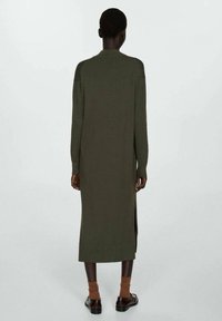 Personne vue de dos portant une longue robe-pull verte olive, des chaussettes marron et des chaussures en cuir marron foncé sur un fond clair uni.