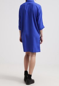 Loose-fitting, solid blue shirt dress with a collar and rolled-up sleeves. Smooth texture, falls above the knees, paired with black footwear.