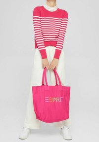 Osoba w białych spodniach i białych trampkach trzyma jaskraworóżową torbę tote z wielokolorowym napisem "ESPRIT" oraz swetrem w czerwono-białe paski.