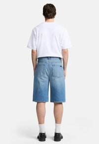 Shorts en denim délavé, coupe décontractée, longueur genou, avec deux poches arrière, associés à un t-shirt blanc uni et des chaussures noires.