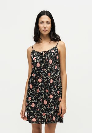 Vrouw met donker haar tot op de schouders, die een zwarte mouwloze jurk met bloemenprint en dunne bandjes draagt tegen een effen lichte achtergrond.
