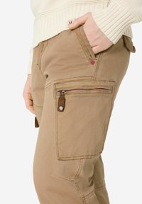 Beige Cargohosen mit strukturierter Oberfläche, ausgestattet mit mehreren Taschen, einer seitlichen Reißverschlusstasche und roten Akzentnähten.