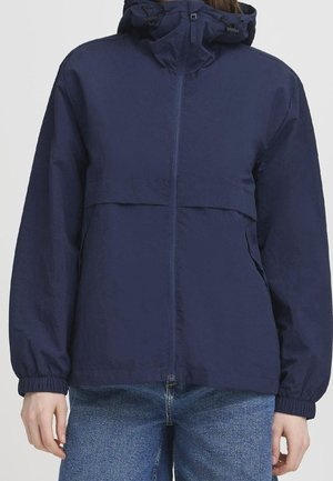 Osoba ubrana w granatową kurtkę z kapturem z przednim zamkiem błyskawicznym i elastycznymi mankietami, w połączeniu z niebieskimi dżinsami, stojąca na tle jednolitego tła.