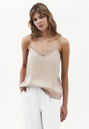Vrouw met lichtbruin golvend haar, die een beige hemdje met kanten rand en witte hoge taille broek draagt tegen een witte achtergrond.