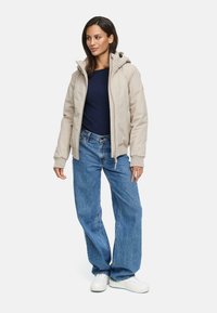 Lichtbeige geïsoleerde jas met een capuchon, ritsluiting en zijzakken over een marineblauw longsleeve shirt. Draag het met losse blauwe jeans en witte sneakers.