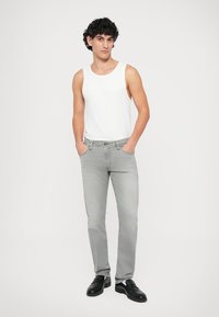 Szürke farmernadrág slim fit szabással, sima fehér topless, és fekete dresszcipő. A farmernadrág öt zsebet tartalmaz és klasszikus gombos záródással rendelkezik.