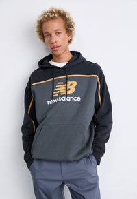 Pelēka kapuce ar melniem piedurknēm, ar dzeltenu un baltu "New Balance" logotipu. Iekļauj priekšējo kabatu un auklu.