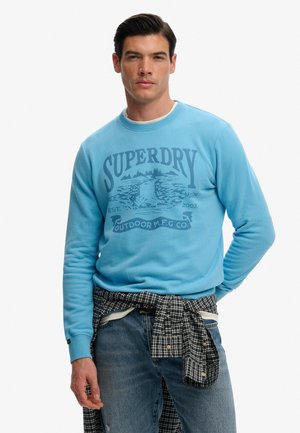 Hombre con sudadera Superdry azul claro, camisa de cuadros atada alrededor de la cintura y vaqueros azules, de pie frente a un fondo blanco.