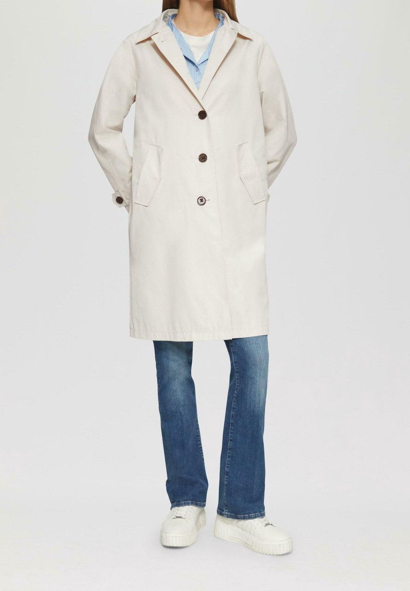 Personne portant un manteau beige mi-long avec des boutons marron, un jean bleu, des baskets blanches et une chemise bleu clair portée sur un haut blanc.