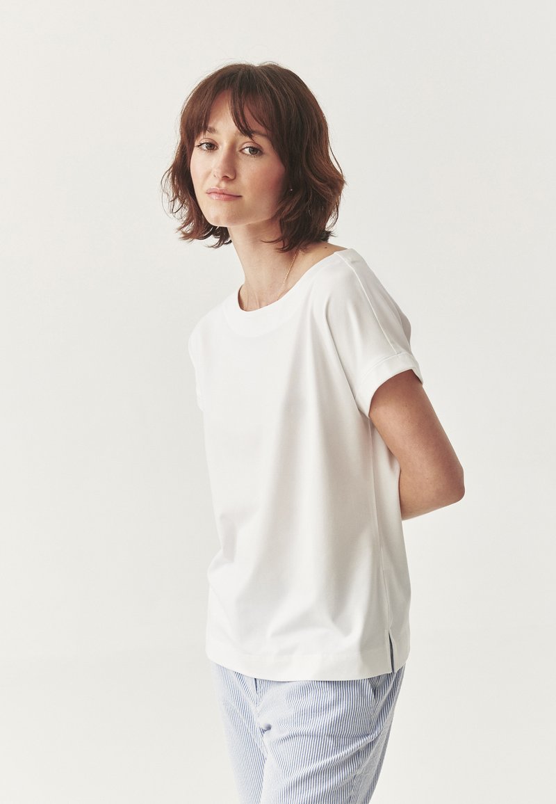 White short-sleeve shirt with a round neckline and relaxed fit, featuring a smooth fabric and side slit hem, paired with striped pants.