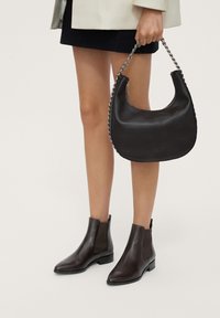 Botas de tobillo de cuero marrón con inserciones laterales elásticas, punta redondeada y un tacón bajo tipo bloque. Se sostiene un bolso de cuero negro con un asa de cadena.