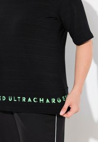 Camiseta negra de manga corta con tela texturizada, que presenta un texto gráfico verde "ULTRACHARGED" a lo largo del dobladillo, combinada con pantalones negros.