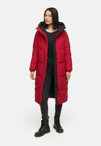 Rød knælængde pufferjakke med hætte, med en sort foring og et firkantet quiltet design. Båret med sorte jeans og snørebåndsstøvler.