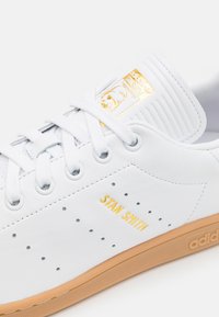 Deportiva blanca de cuero con punta redonda, acentos perforados, logo dorado "STAN SMITH" y suela de goma. Presenta cierre con cordones y marca en la lengüeta.