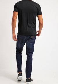 Camiseta negra de manga corta combinada con jeans de mezclilla azul oscuro. Los jeans presentan un lavado sutil y detalles en los bolsillos traseros. Zapatillas negras completan el look.