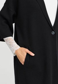 Black cardigan with a soft texture, featuring a v-neck, two pockets, and lace accents on the cuffs. Fastened with a single button.