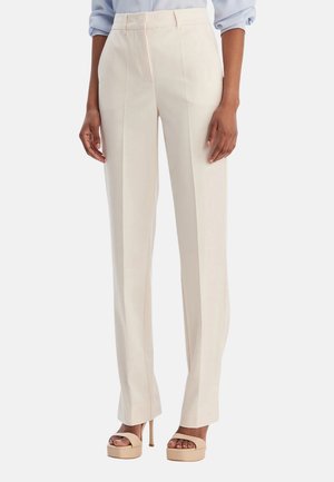 Donna che indossa pantaloni su misura a vita alta color crema con pieghe frontali e sandali beige aperti in punta con tacco alto, in piedi con le mani rilassate.