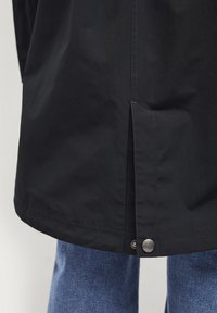 Manteau noir imperméable avec un ourlet asymétrique, fente latérale et fermeture à pression métallique. Texture lisse avec des détails de couture visibles.