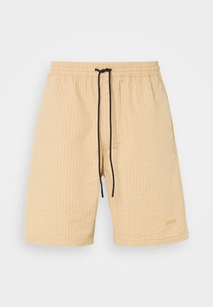 Beige shorts with a textured fabric, elastic waistband, and black drawstring. Features a subtle logo accent. Straight-cut design.
