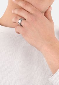 Un primo piano di una mano con un anello d'argento che tocca delicatamente il mento di una persona che indossa una camicia bianca a trama.