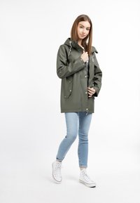 Pelerină de raincoat verde, impermeabilă, cu glugă, prevăzută cu nasturi pe față și două buzunare laterale, combinată cu blugi albaștri deschiși și sneakers albi.