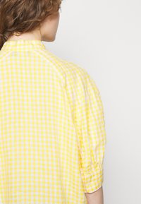 Femme aux cheveux bouclés bruns portant une chemise ample à carreaux jaunes et blancs, vue de dos sur un fond clair uni.