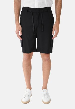 Uomo che indossa pantaloncini cargo neri con coulisse in vita, sneakers bianche e una camicia bianca a maniche corte, in piedi davanti a uno sfondo semplice.
