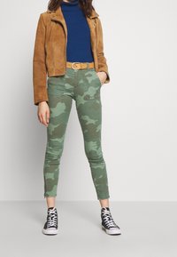 Mujer de pie con una mano en el bolsillo, vistiendo una chaqueta de ante marrón, un suéter de cuello alto azul, pantalones de camuflaje verde y zapatillas negras de caña alta.