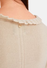Beige gestrickter Pullover mit einem gerippten Kragen und einem rüschenden Saum, ausgestattet mit einem metallischen Knopfverschluss auf der Rückseite und einem strukturierten Muster.