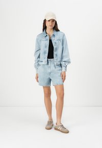 Veste en denim bleu clair et shorts, haut noir, casquette beige texturée et baskets grises avec des accents roses. Tenue décontractée sur un fond uni.