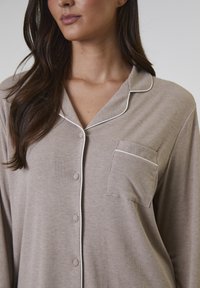 Beige geribbelde pyjama-top met witte piping, knoopsluiting aan de voorkant en een borstzak. Gladde stof met een relaxte pasvorm en kraagontwerp.