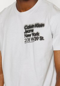 Bílý bavlněný triko s černě potištěným logem a textem „Calvin Klein Jeans New York 205 W39 St.“ na přední straně. Krátké rukávy, kulatý výstřih.
