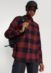 Camisa de flanela xadrez vermelha e azul-marinho com dois bolsos no peito, fechos de botão e mangas compridas, combinada com uma mochila preta e uma chapéu.