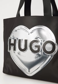 Melna auduma soma ar sudraba sirds grafiku, uz kura ir uzraksts "HUGO" izteiksmīgajās melnajā burtiem. Dizainam ir gluda, spīdīga tekstūra.