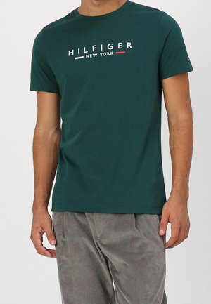 Homme portant un T-shirt à manches courtes vert foncé avec le texte "HILFIGER NEW YORK", associé à un pantalon en velours côtelé gris clair.