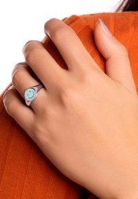 Anillo de plata con un diseño de cara sonriente de color azul claro, llevado en una mano sobre un fondo de tela ribeteada naranja. Textura metálica suave.