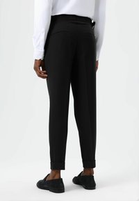 Pantalon noir sur mesure à coupe slim, avec ourlets retroussés, deux poches arrière et texture lisse, porté avec une chemise blanche à manches longues.