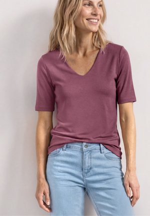 Kvinde med blondt hår iført en lilla V-hals kortærmet skjorte og lyseblå jeans, smilende og kiggende mod højre.