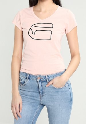 Kobieta w jasnoróżowej koszulce z dekoltem w serek ozdobionej czarnym abstrakcyjnym wzorem oraz jasnoniebieskich dżinsach, z jedną ręką w kieszeni.