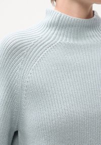 Pull en maille côtelée bleu clair avec col montant porté par une personne, montrant la texture détaillée et les motifs de points sur l'épaule et la poitrine.