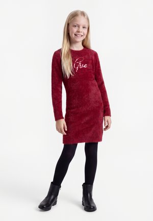 Jeune fille souriante, portant une robe rouge duveteuse à manches longues, des collants noirs et des bottines noires, debout devant un fond uni.
