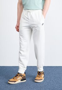 Pantaloni albi din bumbac cu talie reglabilă, buzunare laterale și un mic logo Nike de culoare neagră. Tenisi înalți maro și bej cu accente.