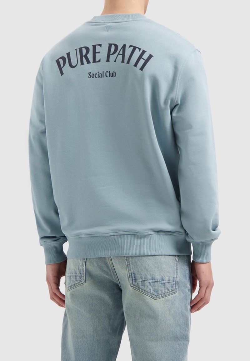 Light blue sweatshirt with ribbed cuffs and hem. Back features "PURE PATH Social Club" in dark blue curved text. Soft fabric texture.